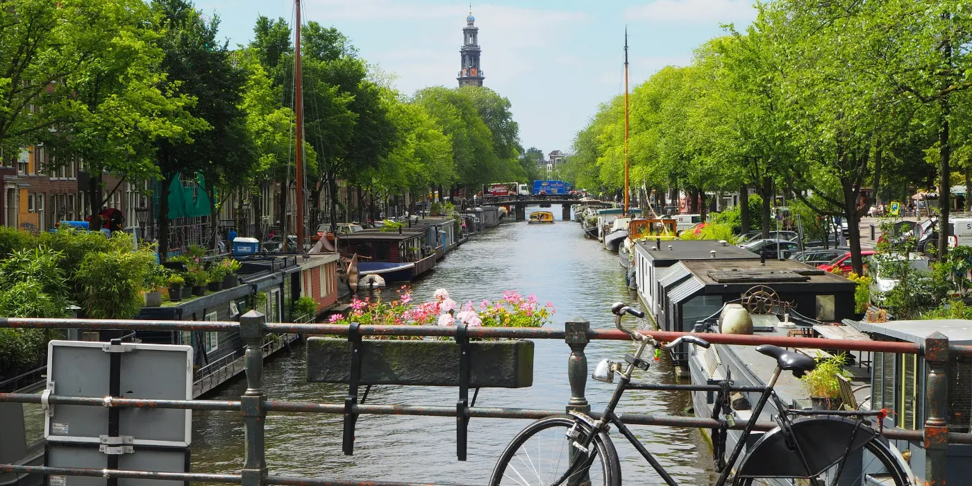 A canal in Amsterdam seen from a bridge, with houseboats moored along both banks, a bicycle leaning against the railing and the Westerkerk tower at the far end.