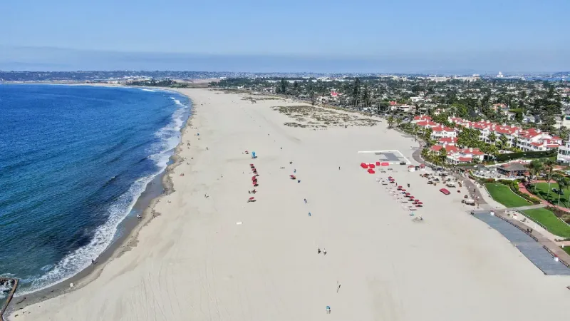 Coronado Central Beach from above, an unusually wide flat expanse of pale sand with the red-roofed hotel beside it.