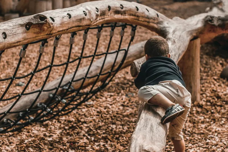 A young child in a dark hooded top climbing over a low log on a wooden playground frame, with a black rope net behind and wood chips underfoot.