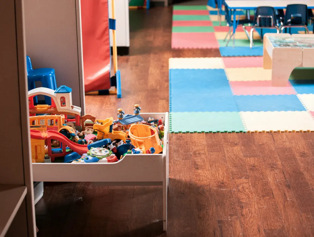 An empty indoor play area, with a low tray of toys in the foreground and interlocking foam floor tiles and small tables beyond it.
